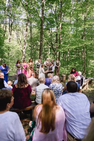 Outdoor forest wedding ceremony with bridesmaids, groomsmen, and guests seated on wooden benches among tall green trees.