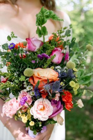 Bride holding a vibrant wildflower wedding bouquet with roses, thistles, and greenery in pink, orange, and purple hues.