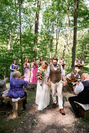 Bride and groom walk down forest aisle holding hands after outdoor woodland wedding ceremony with guests and guitarist.
