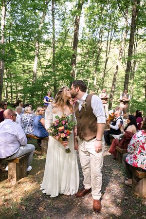 Bride and groom share a kiss walking down outdoor woodland wedding aisle, guests seated on wooden benches among tall trees.