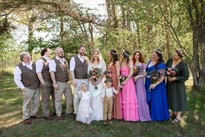 Wedding party poses outdoors in forest setting, bride in white, groomsmen in brown suits, bridesmaids in colorful dresses.