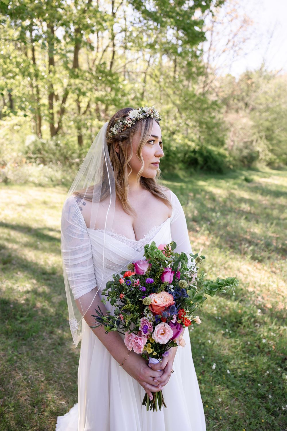 Bride in white gown and floral crown holds colorful bouquet outdoors in lush green forest setting.