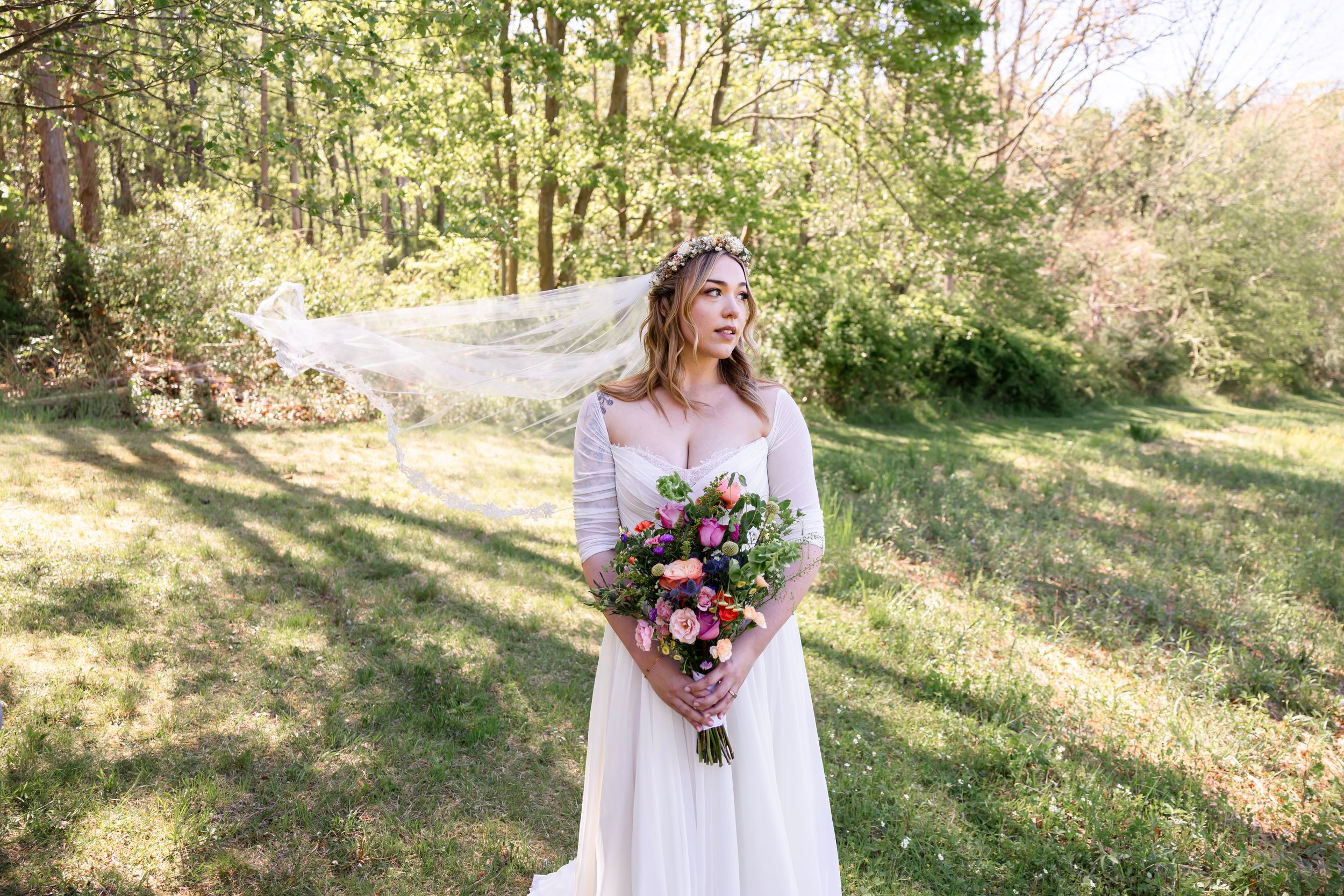 Bride in white gown holding pink bouquet with veil flowing in wind, standing in sunlit forest clearing.