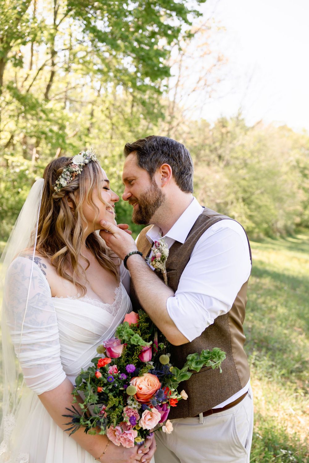 Groom tenderly touches brides face as they share romantic moment outdoors holding colorful boho wedding bouquet.