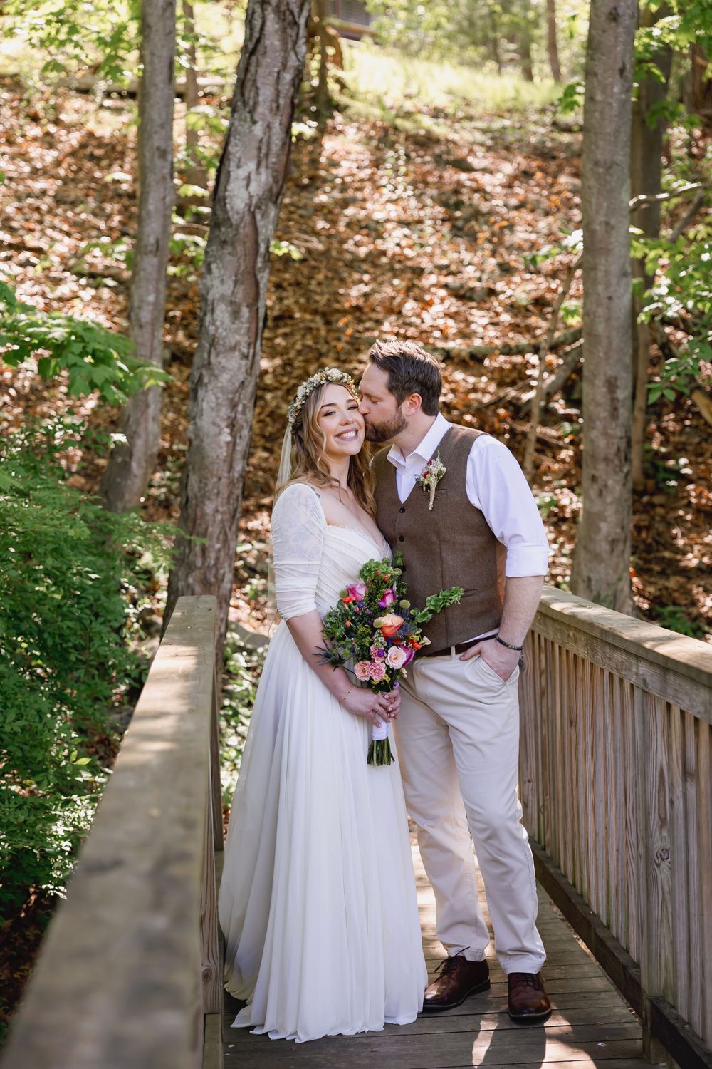 A bride and groom share a romantic kiss on a wooden bridge surrounded by lush forest trees on their wedding day.