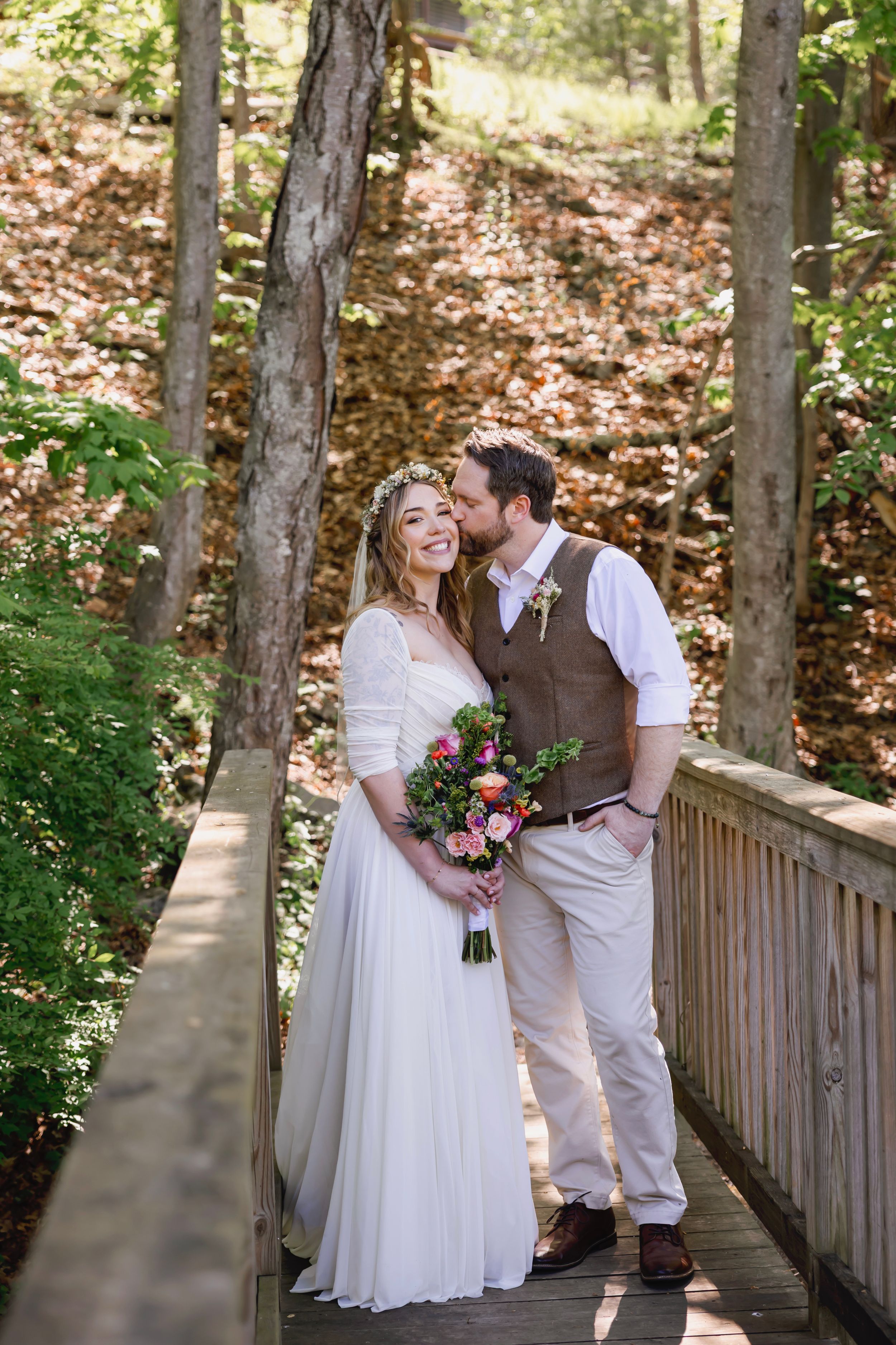 A bride and groom share a romantic kiss on a wooden bridge surrounded by lush forest trees on their wedding day.
