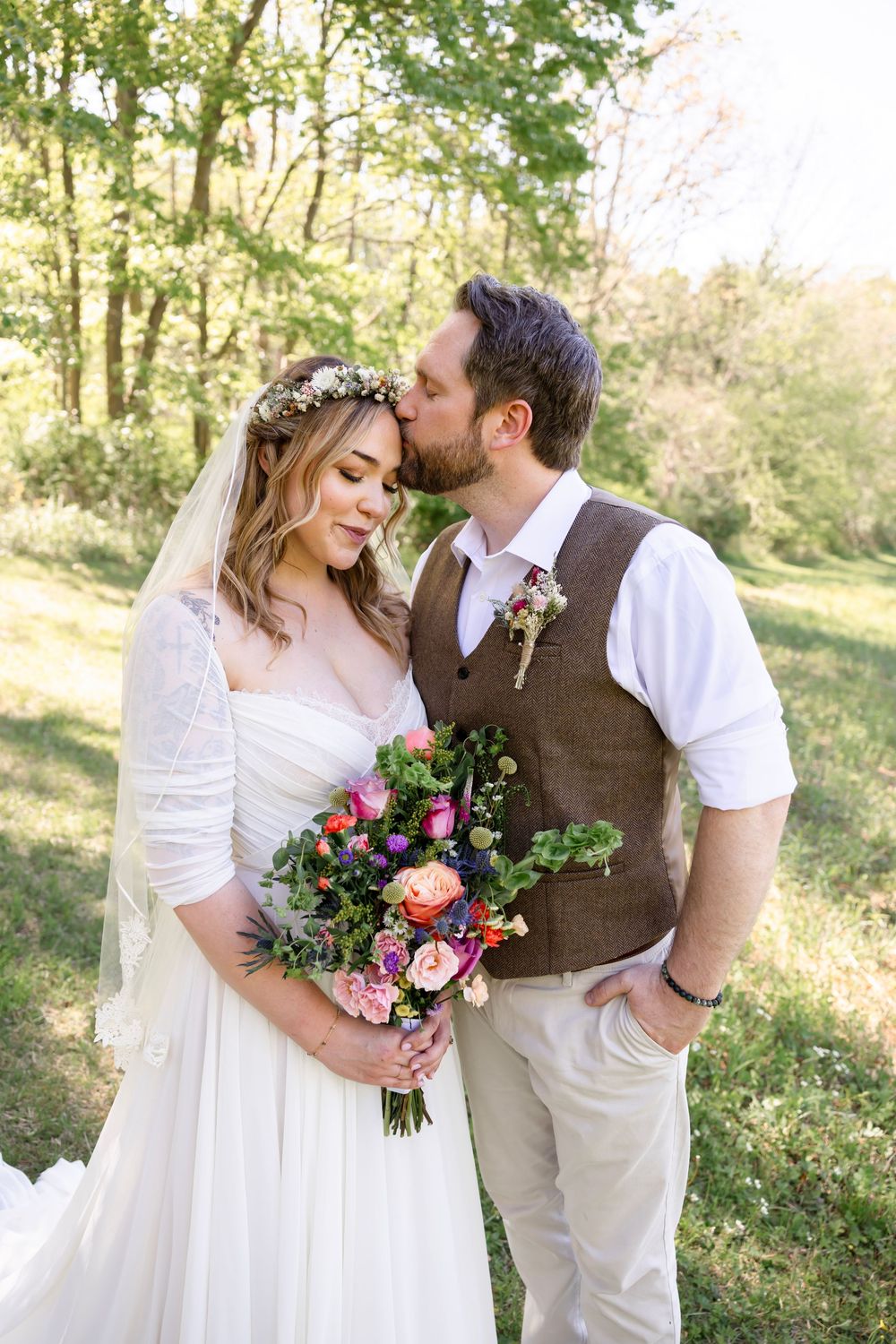 Groom kisses bride on forehead in sunlit forest setting as she holds vibrant colorful bohemian floral bouquet.