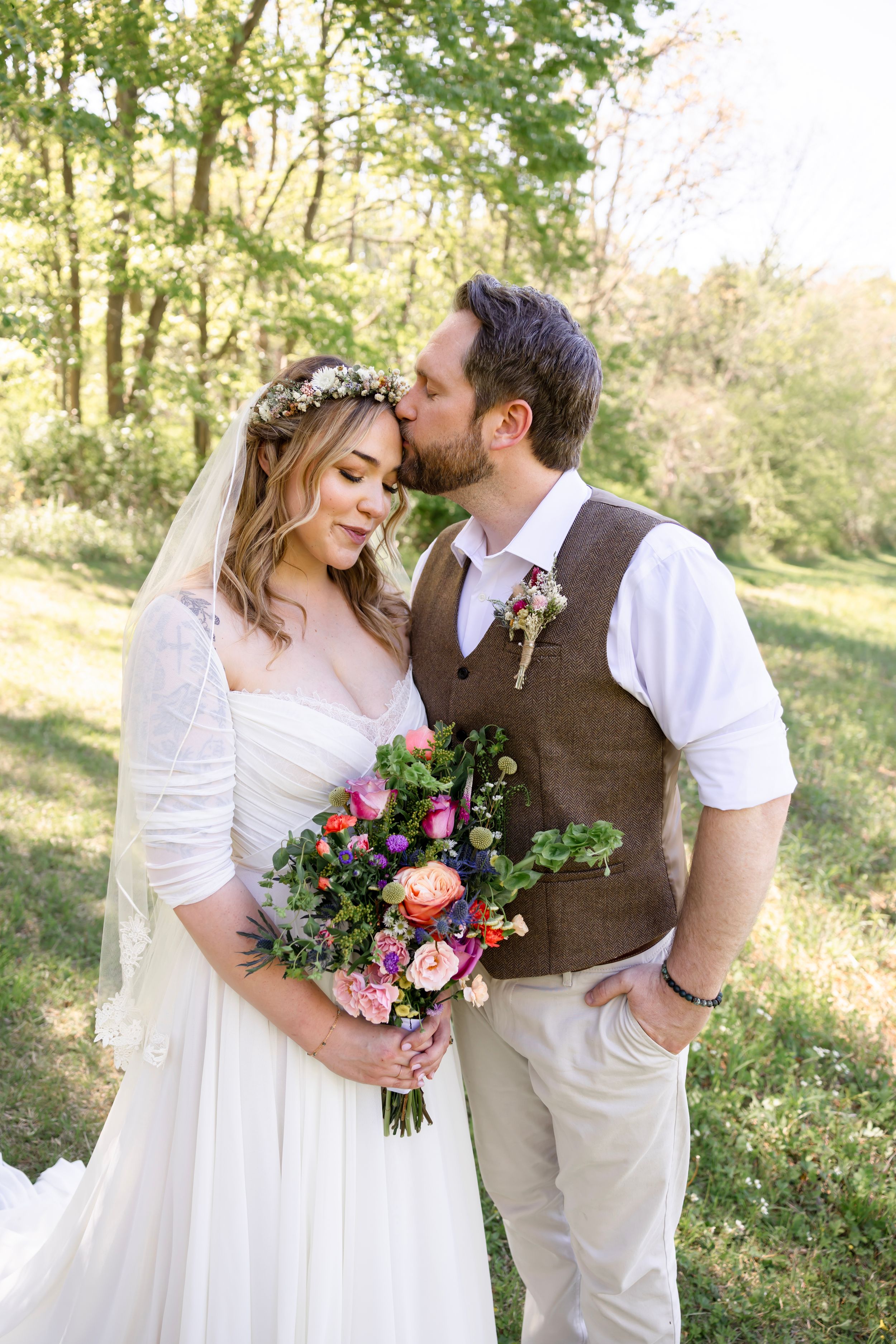 Groom kisses bride on forehead in sunlit forest setting as she holds vibrant colorful bohemian floral bouquet.