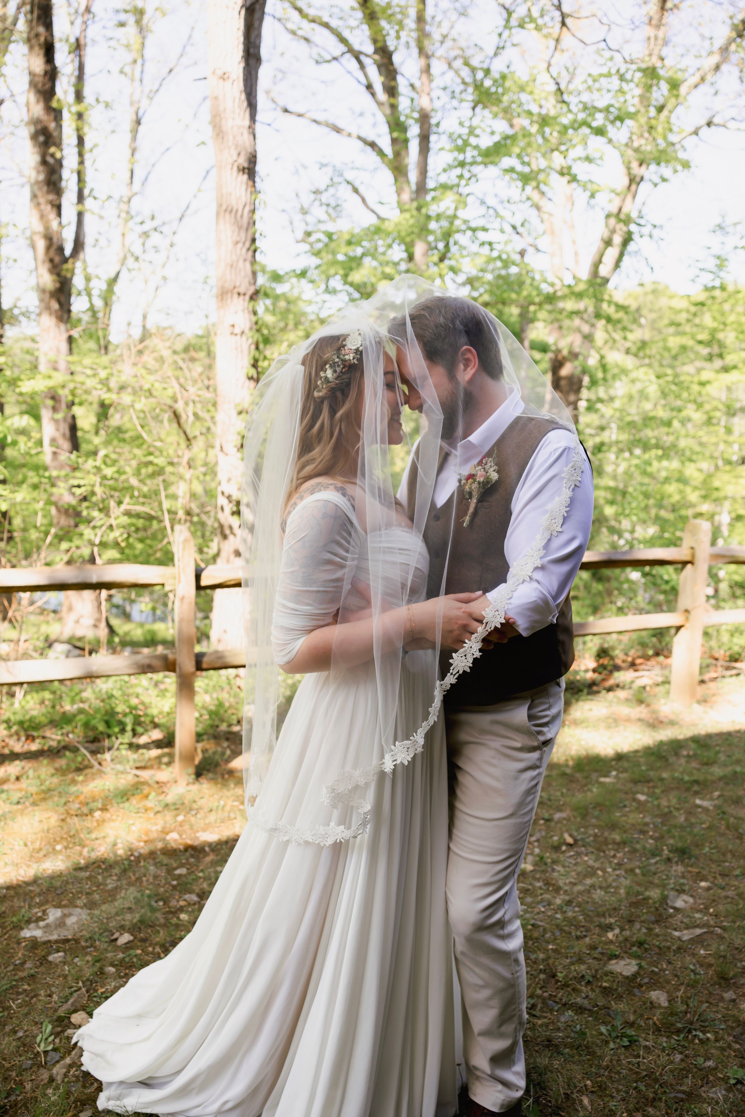 Bride and groom share intimate moment under veil outdoors at rustic woodland wedding ceremony.
