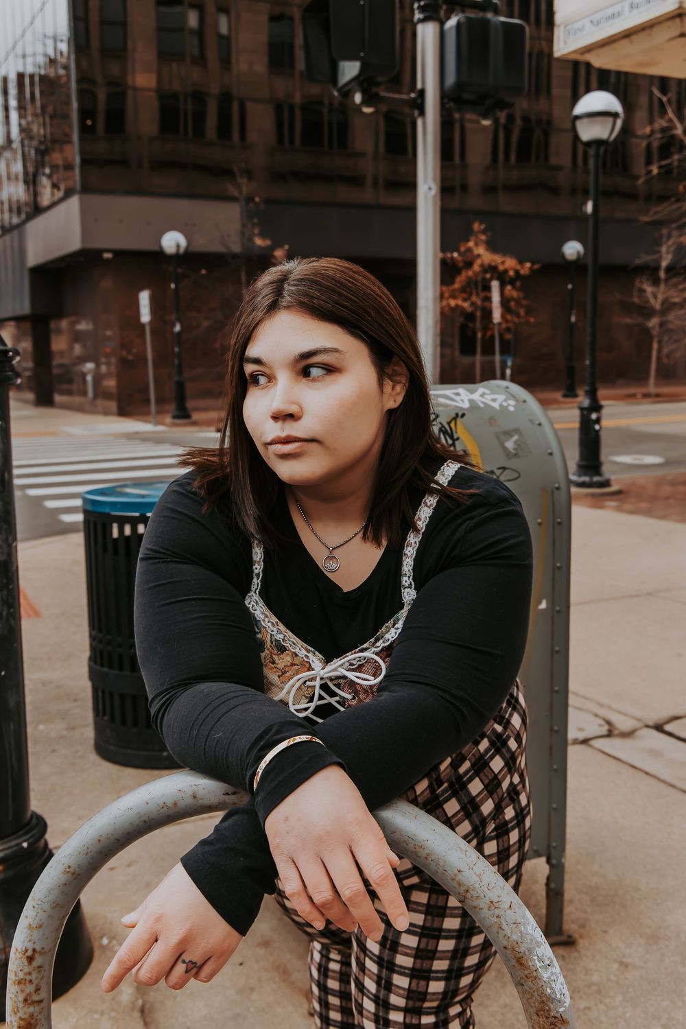 Person wearing a black long sleeve top and checkered pants sits casually on a metal railing on an urban street corner.