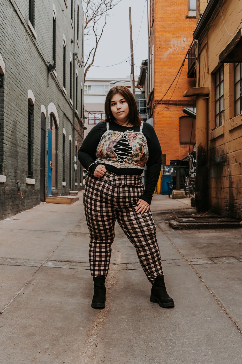 Street style fashion shot featuring black and white plaid pants, graphic top, black cardigan and boots in an urban alley setting.