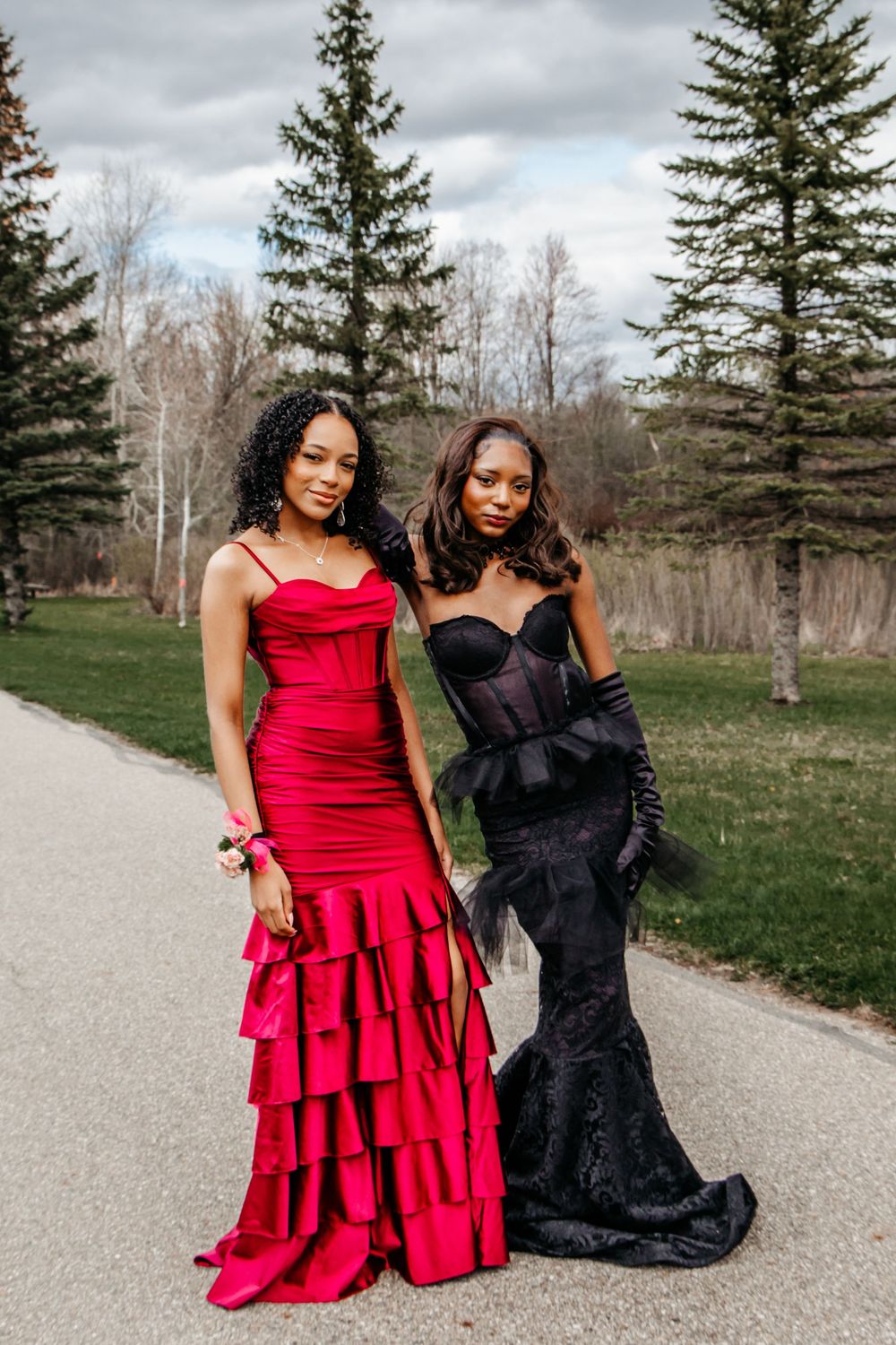 Two friends pose in elegant formal gowns - one in a red ruffled dress and one in a black lace gown with tulle details.