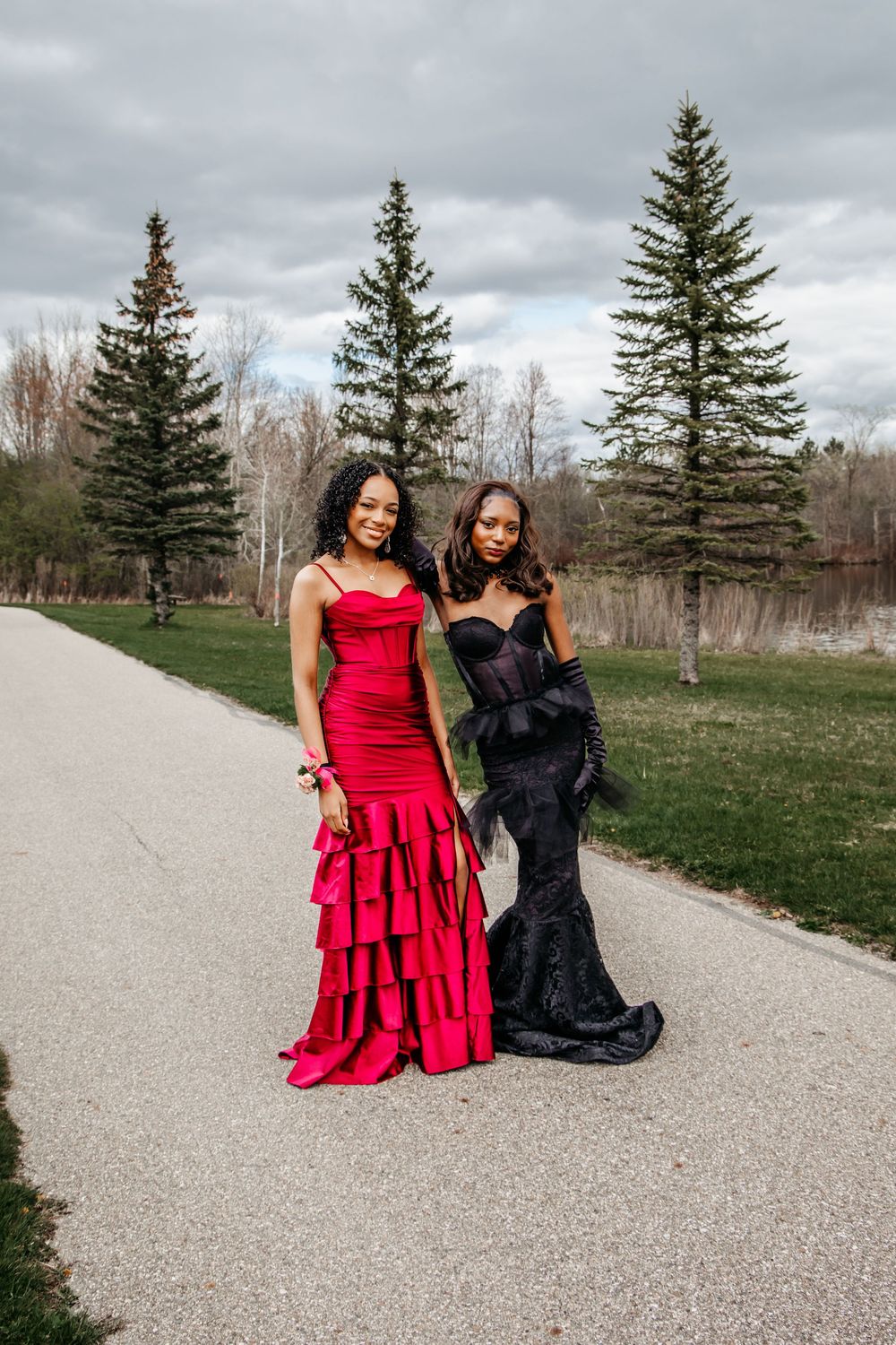 Two models in contrasting red and black formal gowns pose together on a gravel path with evergreen trees in background.