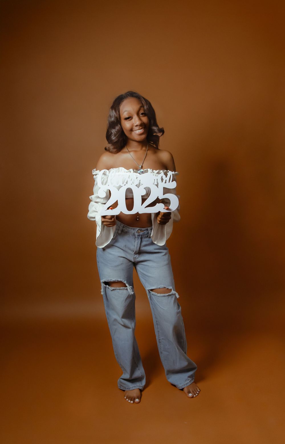 Graduate holds 2025 sign in white off-shoulder top and ripped jeans against brown studio backdrop.