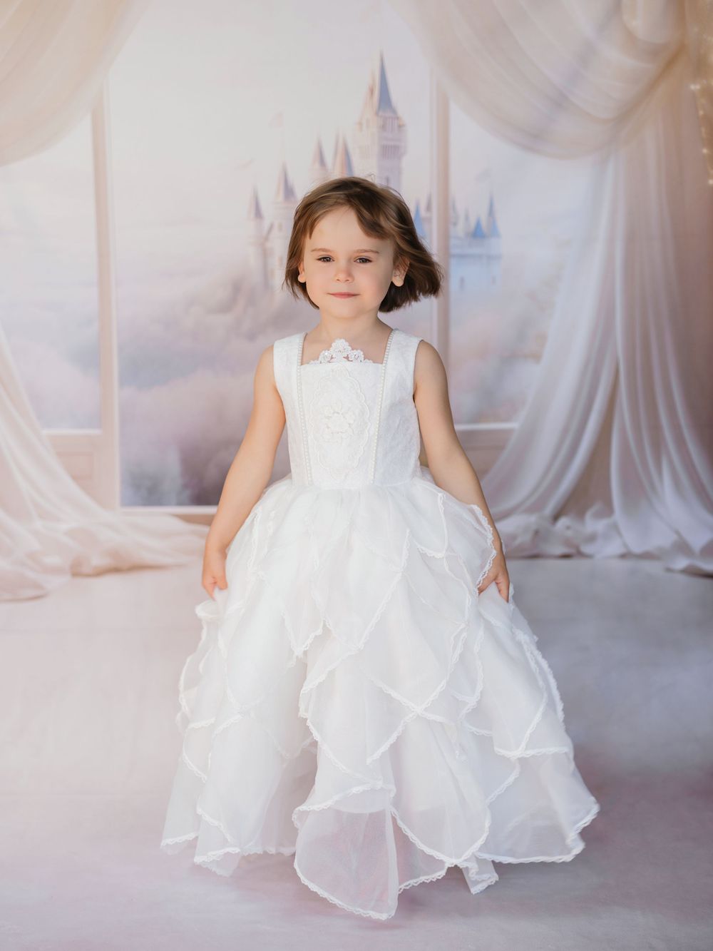 A young child in a white floor-length princess dress poses in front of a dreamy castle backdrop with flowing white curtains.