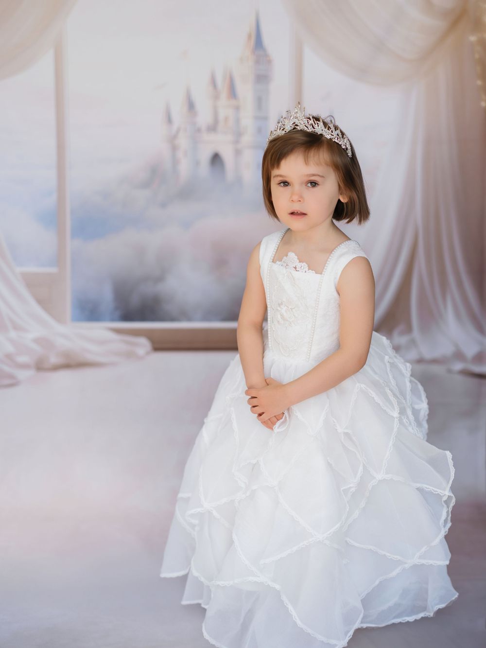 A dreamy princess-themed photo with a white gown, tiara, and ethereal castle backdrop against flowing white fabric.