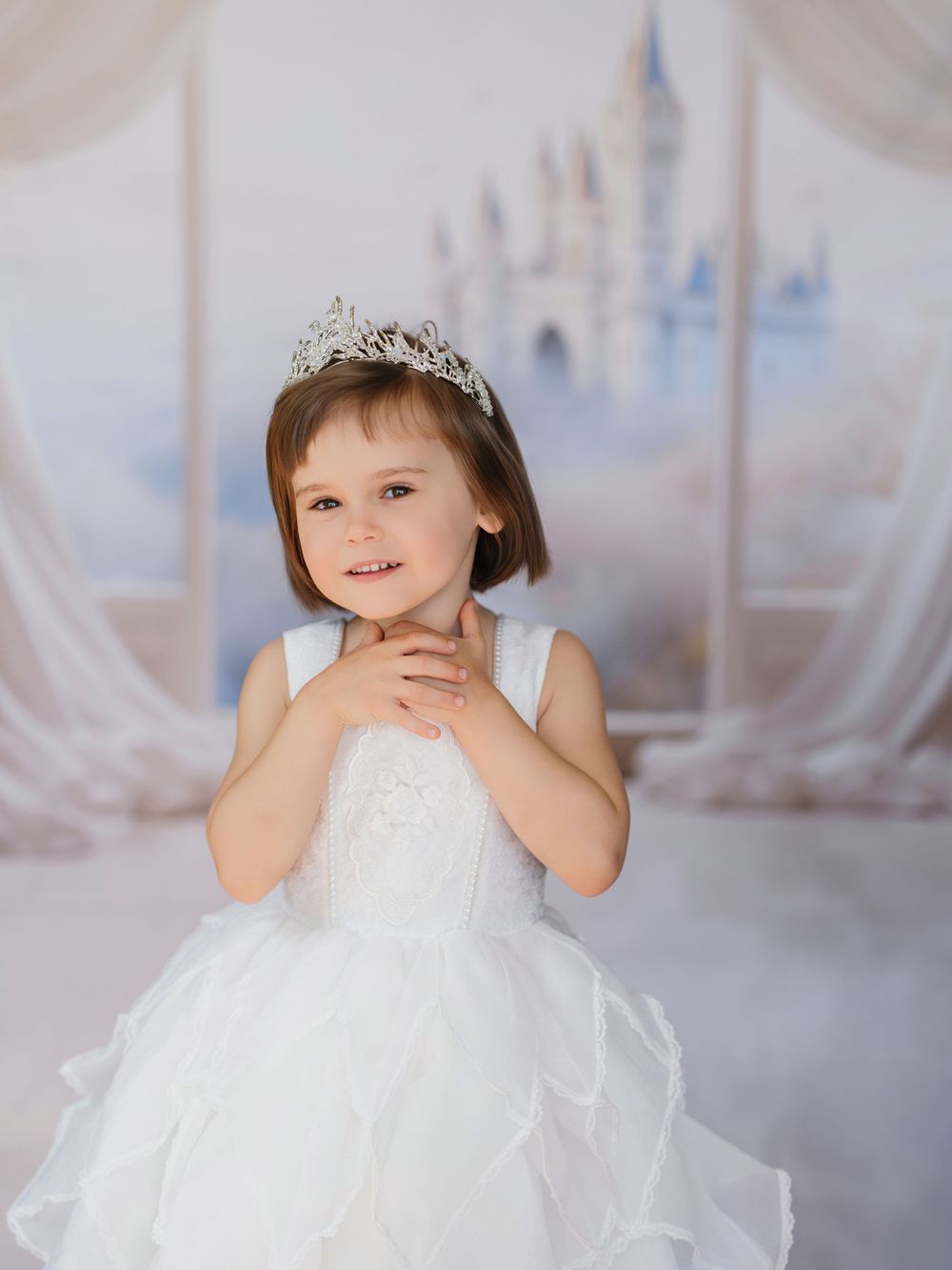 A child wearing a sparkly tiara and white tulle dress poses in front of a dreamy castle backdrop.