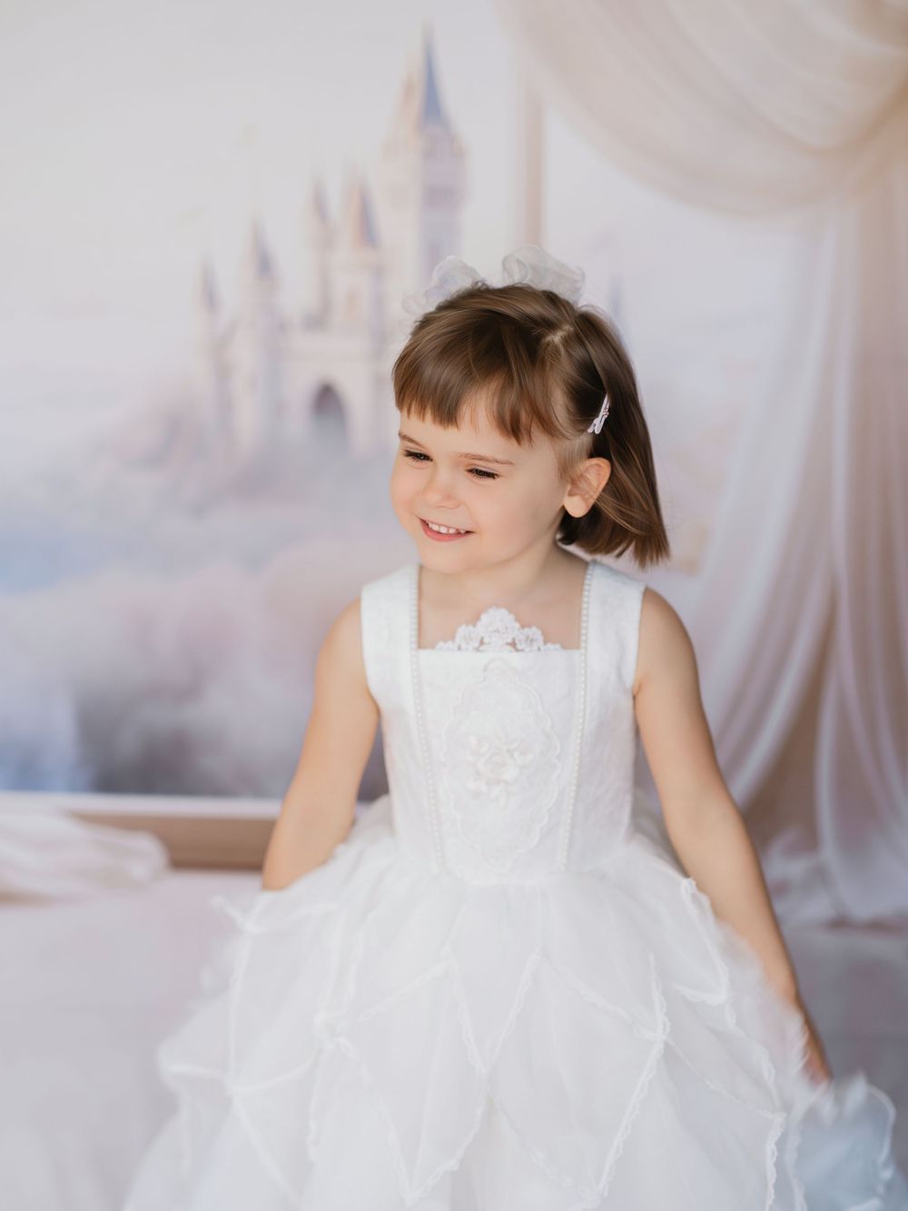 A young child in a white lace dress against a light background twirls and smiles during a portrait session.