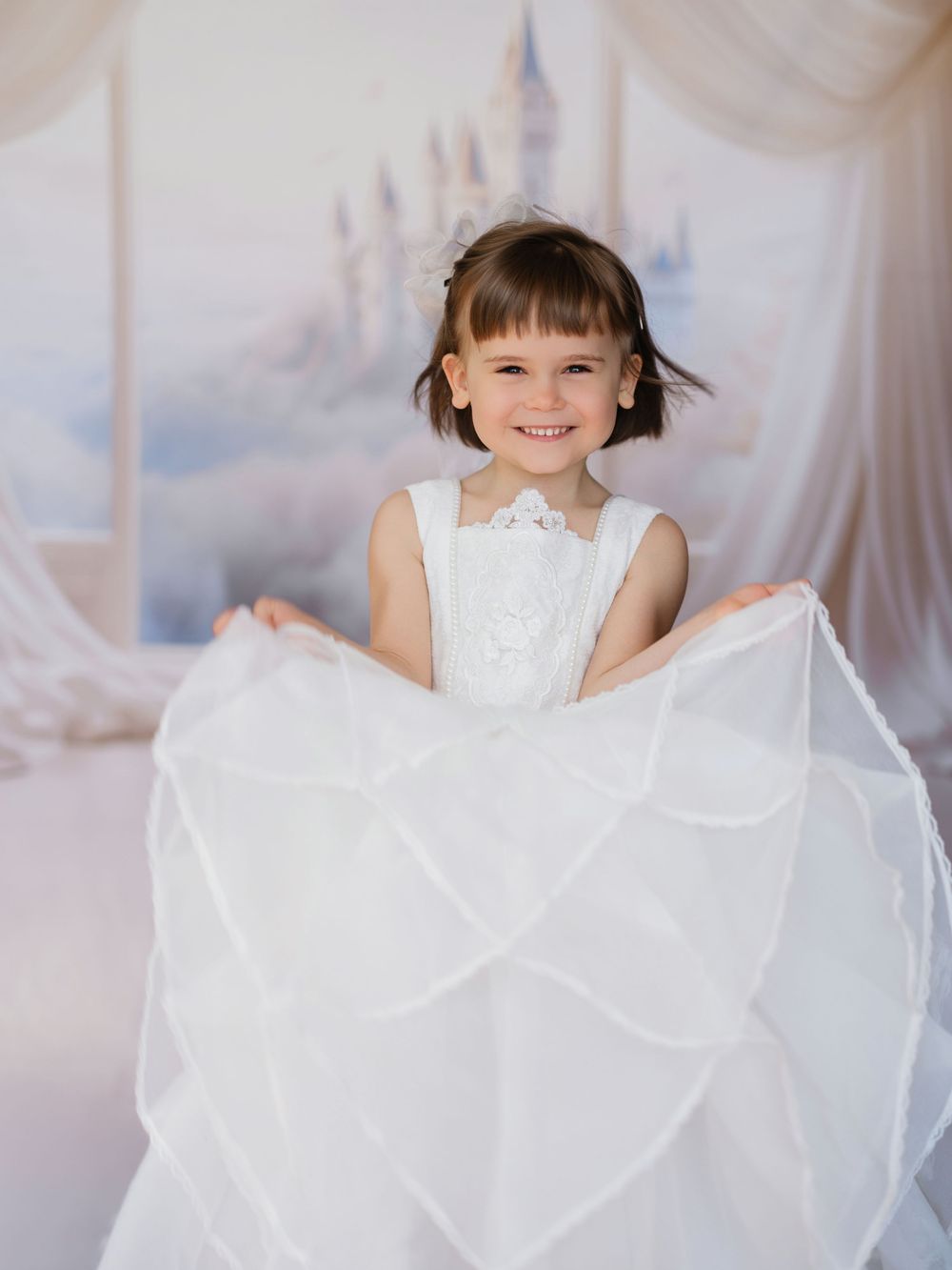 A small child in a flowing white dress twirls joyfully in a dreamy, light-filled studio setting.