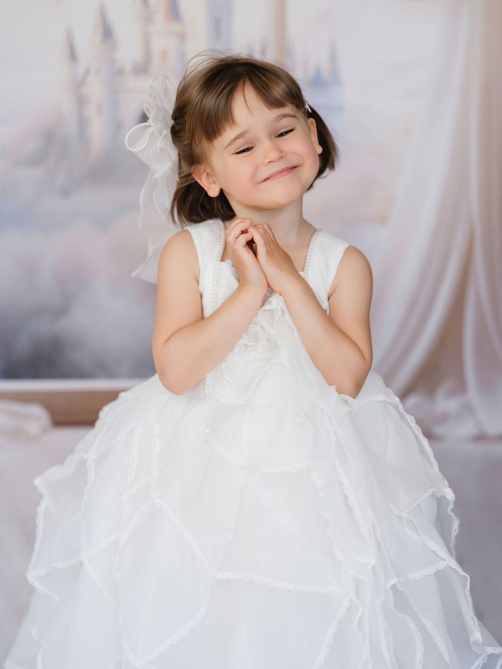 A young child wearing an elegant white frilly dress poses whimsically in a dreamy indoor setting.