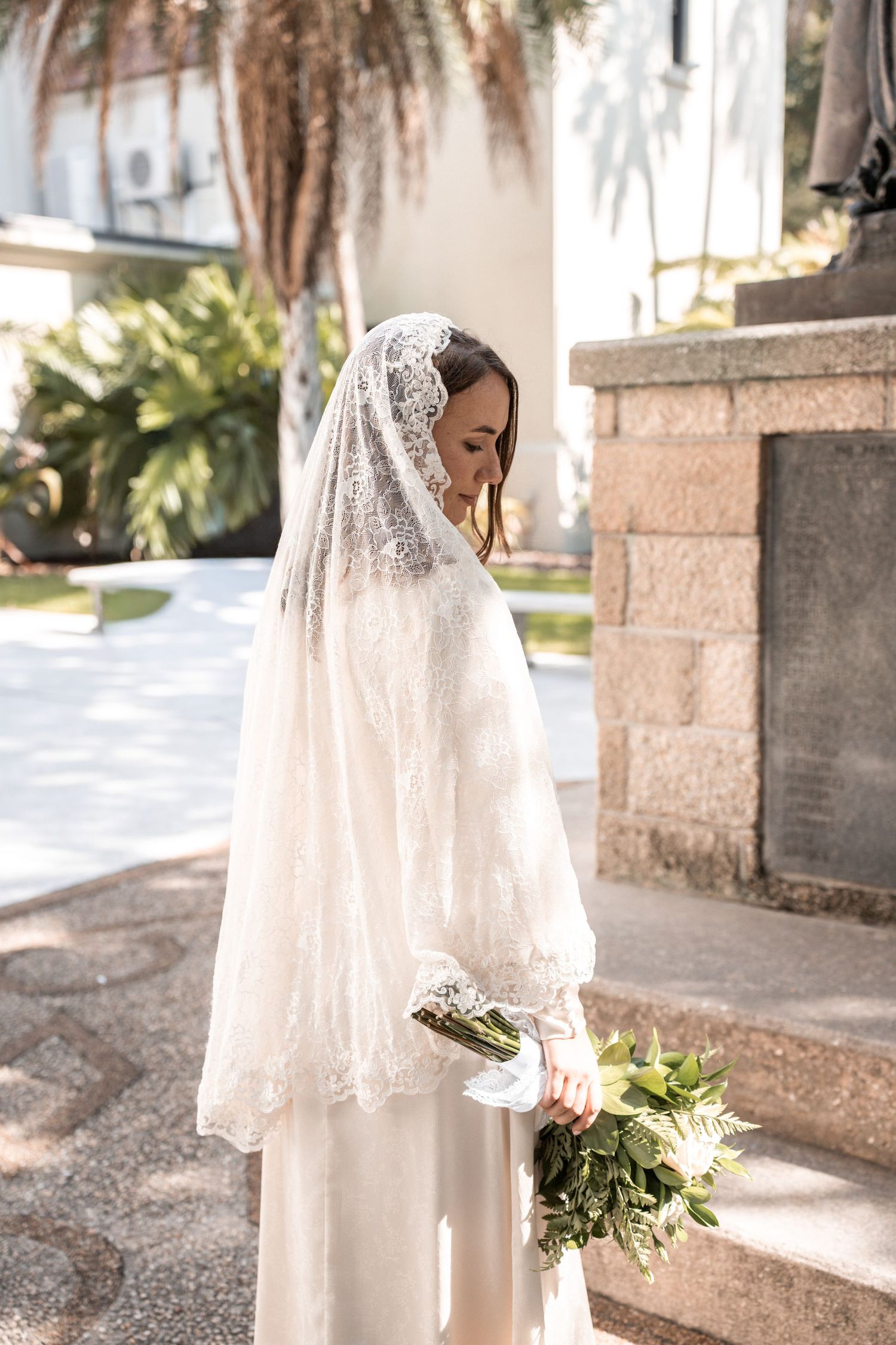 Couple in wedding attire embrace in a garden setting with lush greenery and sunlight filtering through the trees.