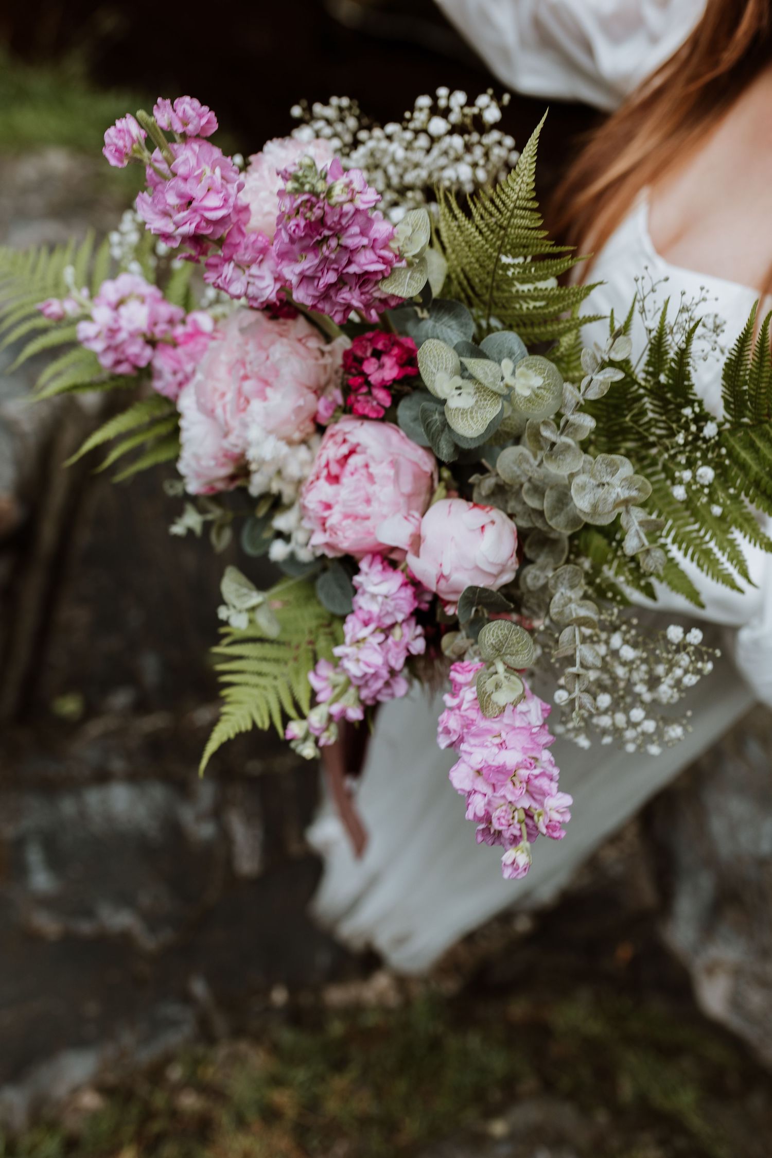 A romantic wedding bouquet featuring pink peonies, stock flowers, white baby's breath and fern fronds in natural light.