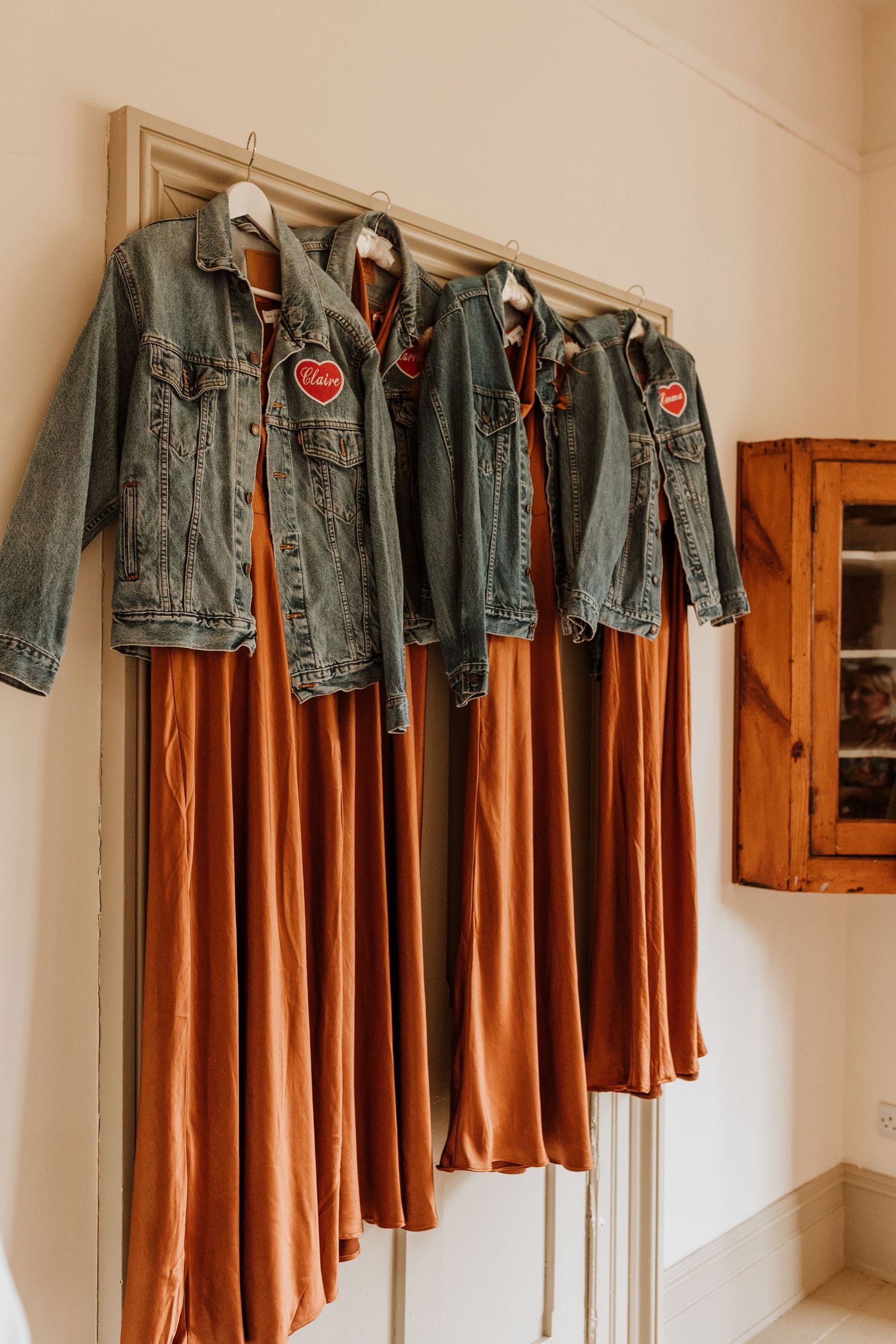 Three denim jackets with red patches hang on hooks beside burnt orange dresses against a beige wall.