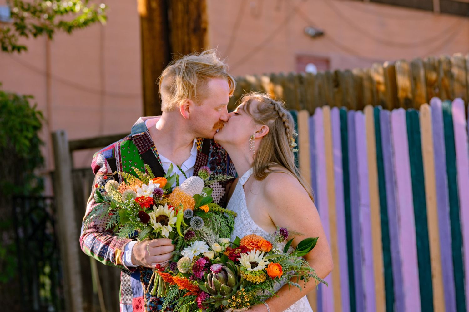 A couple shares a romantic kiss in a garden setting surrounded by vibrant orange and green floral arrangements.
