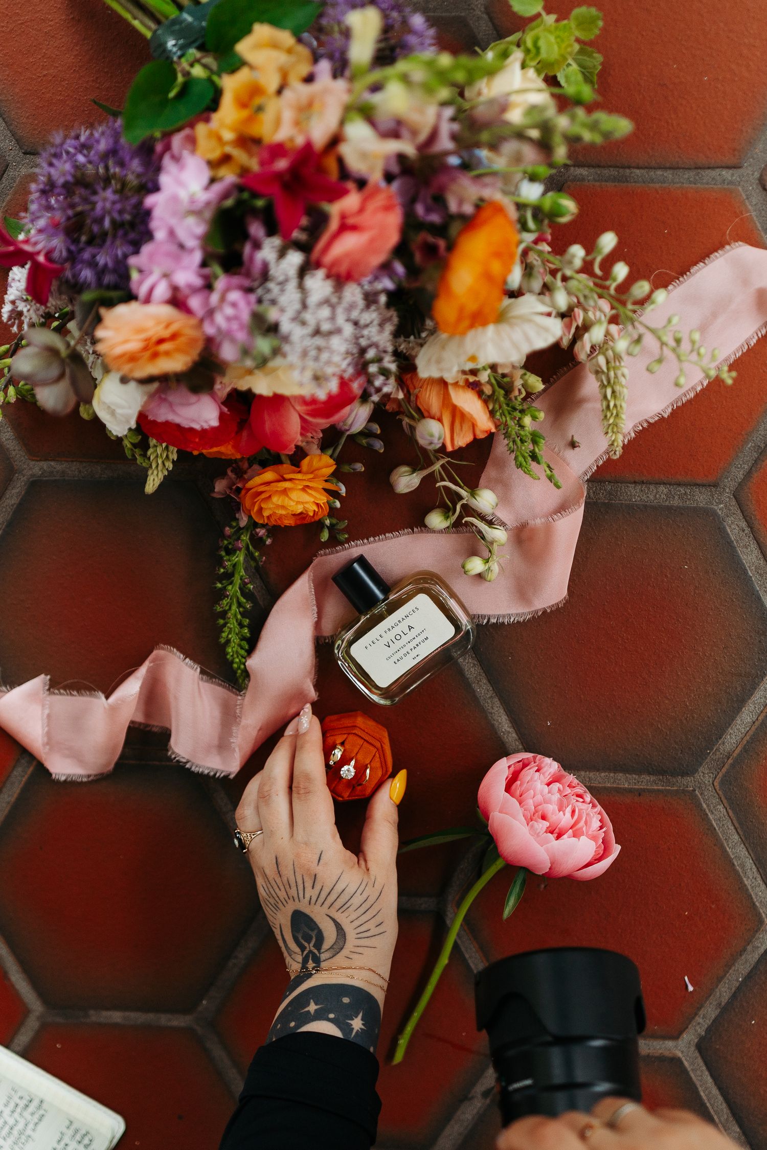 Colorful floral arrangement with perfume bottle on hexagonal tile surface with tattooed hands.