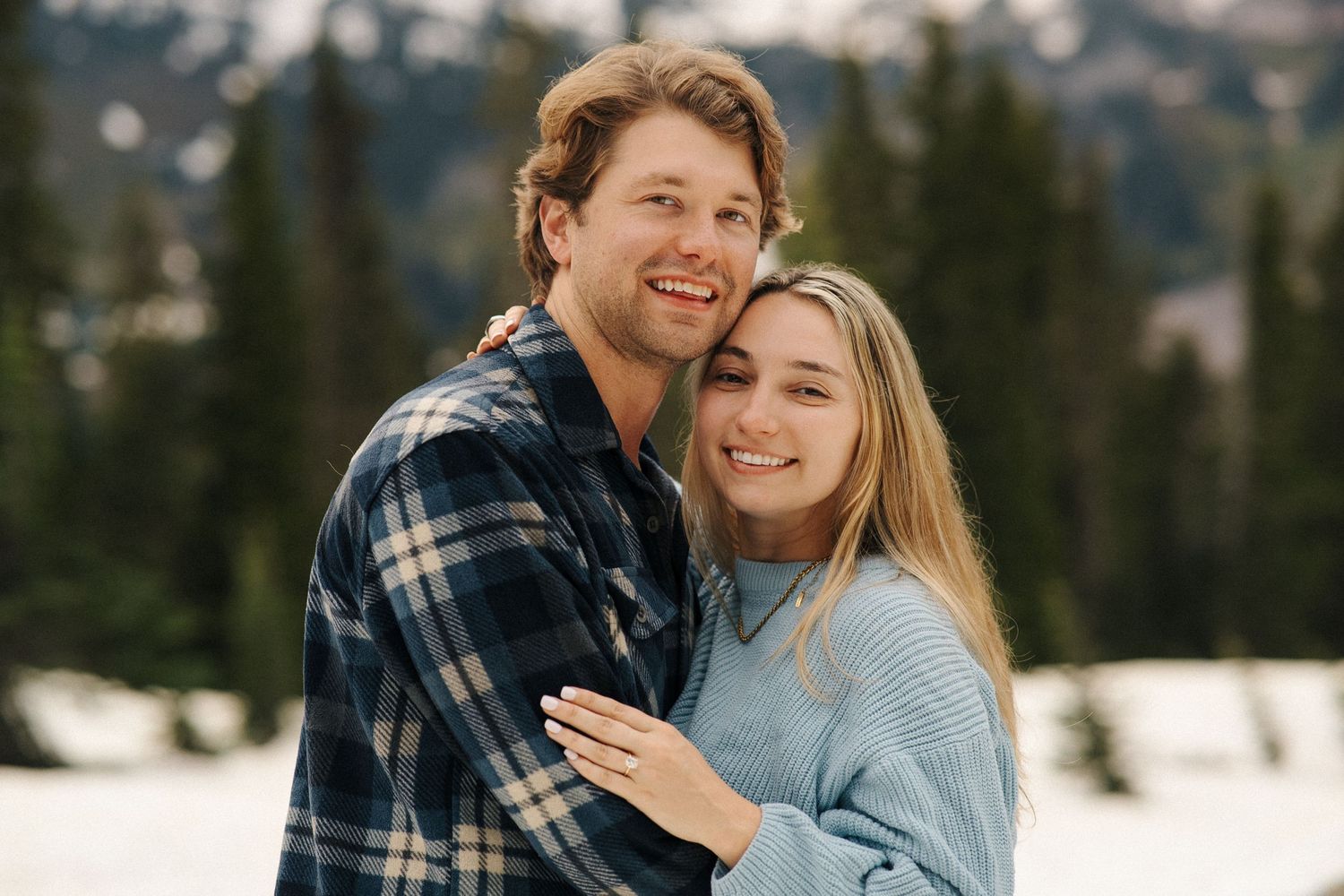 A couple in winter attire embraces and smiles together in a snowy mountain landscape.