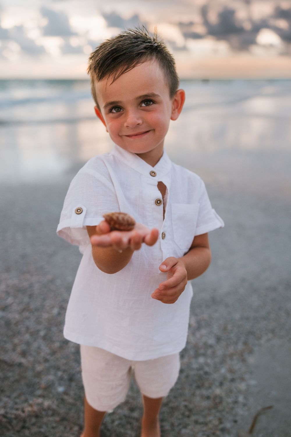 Young child in white linen outfit stands on sandy beach at sunset, reaching out and playing with pebbles.