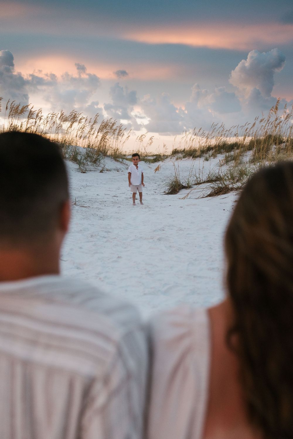 A couple walking along white sandy beach dunes at sunset with sea oats swaying in the foreground.