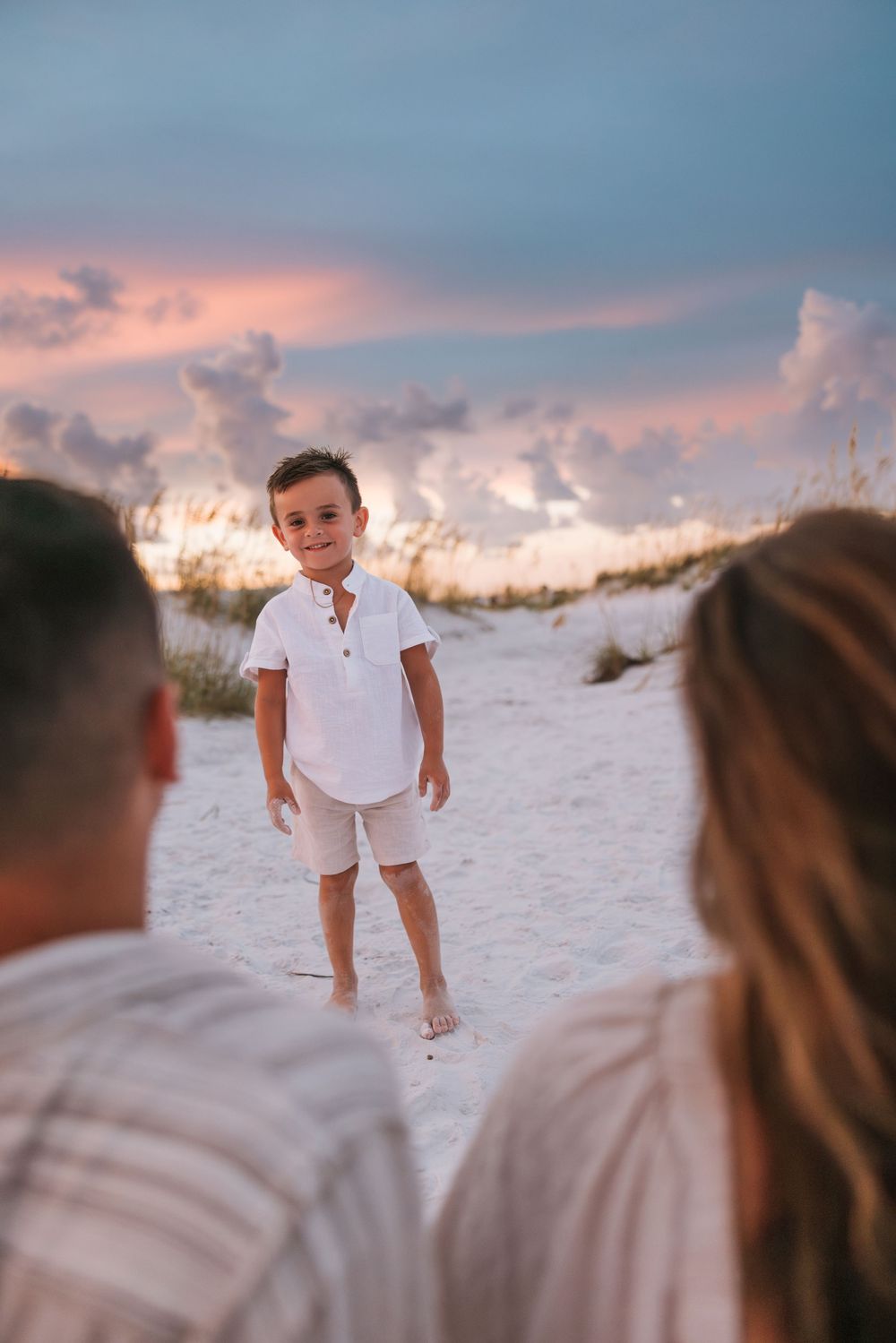 Extended family in white outfits gathers on a sandy beach at sunset with pink cloudy skies.