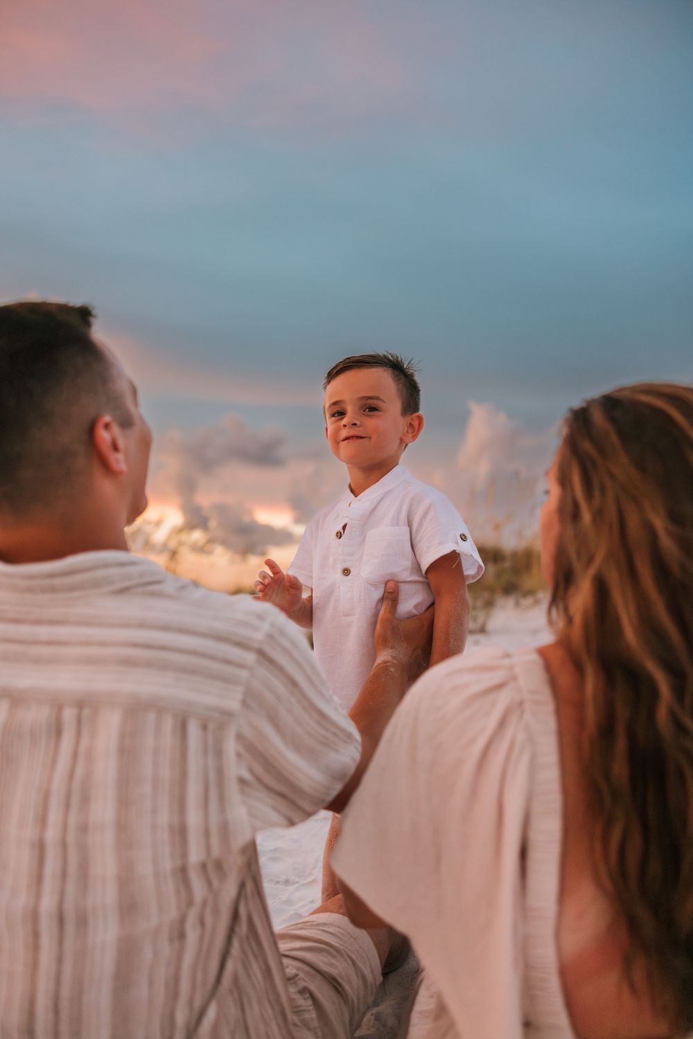 A family in white clothing stands together on a beach at sunset against a tranquil pink and blue sky.