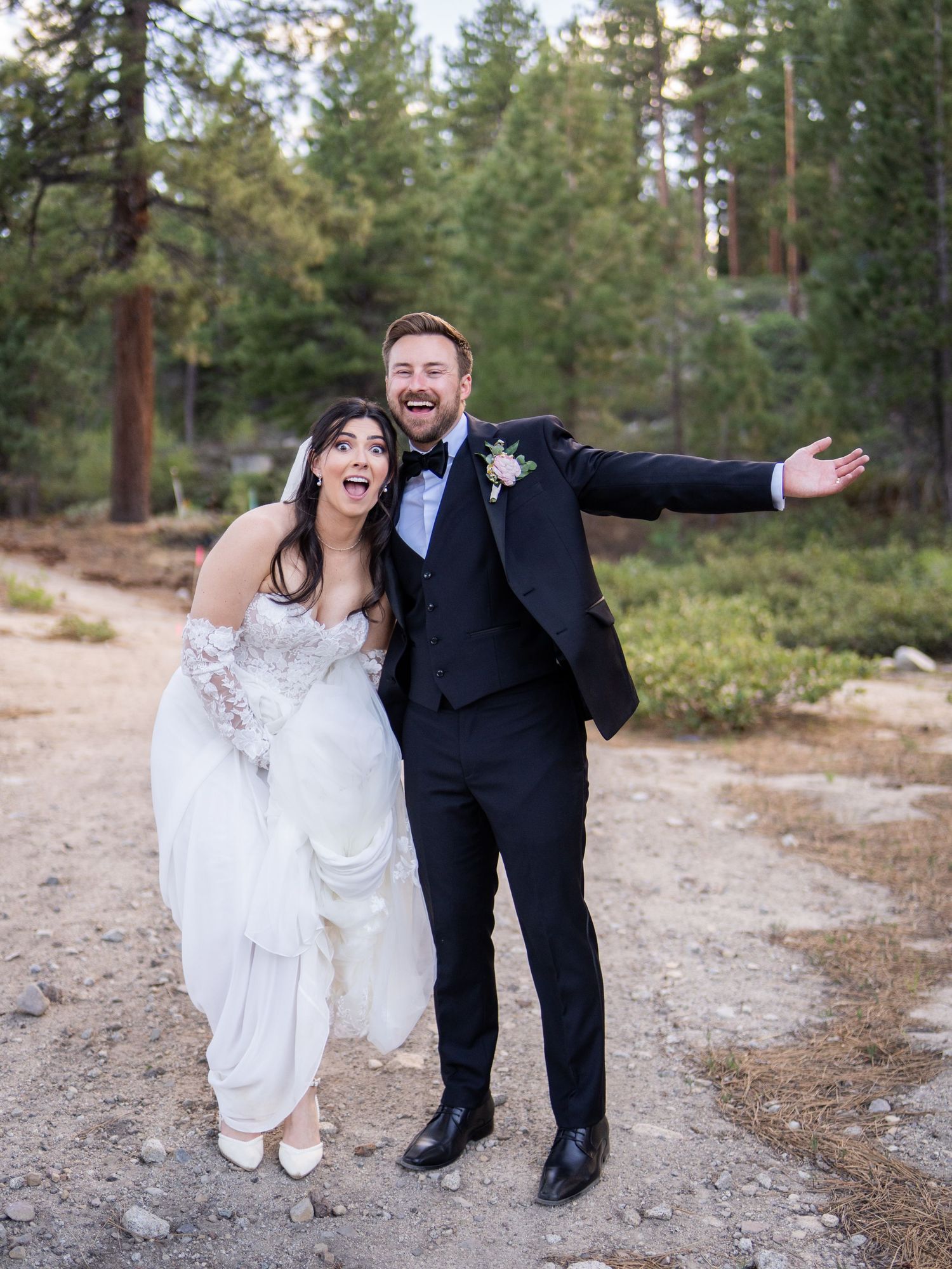 A bride in a white lace wedding dress and veil smiles while holding a white floral bouquet outdoors.