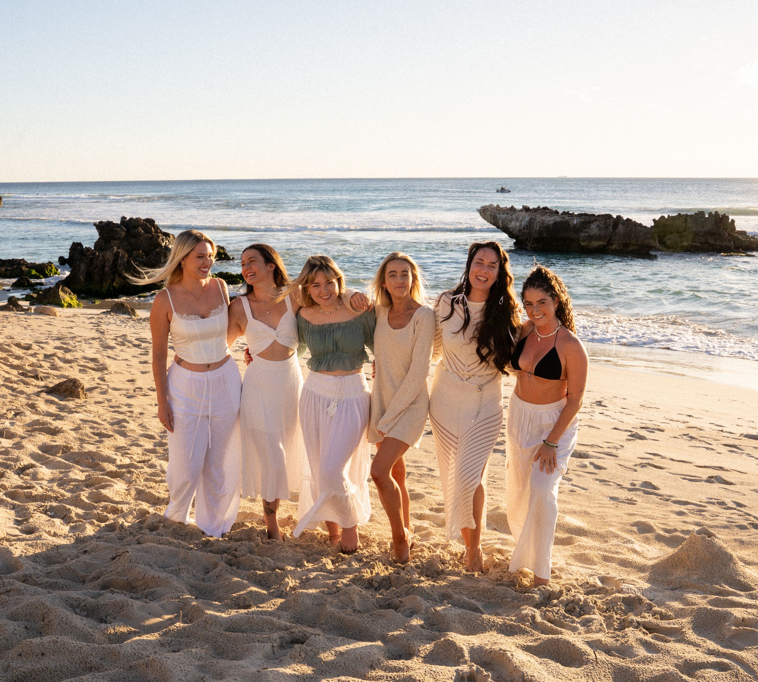 Group of friends in white dresses posing on a sunny beach at sunset.