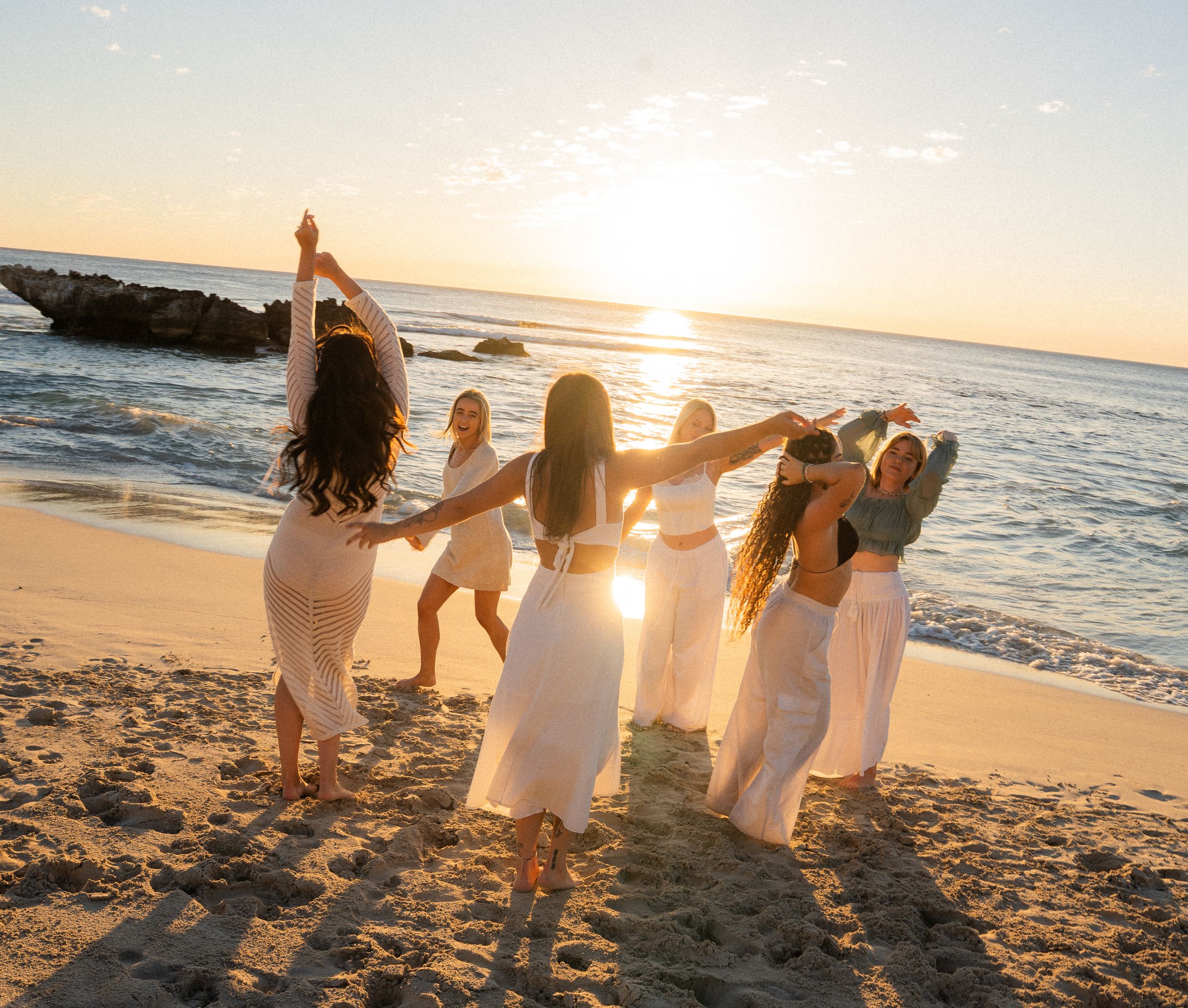Friends wearing white outfits holding hands and walking along the shoreline at sunset.