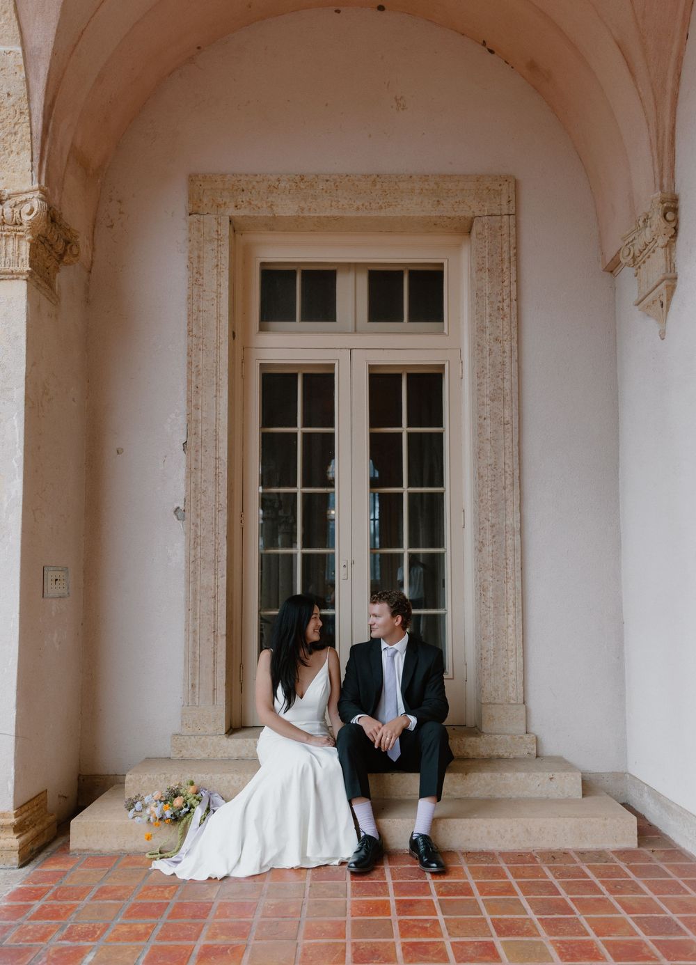 A couple sits together beneath a graceful arched entryway with ornate stone details and terracotta flooring.