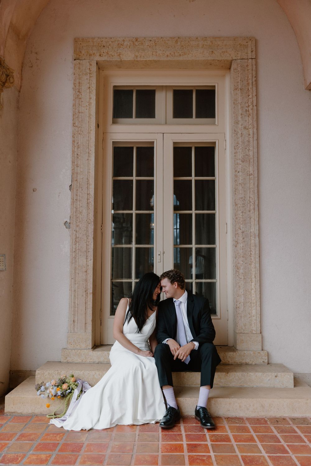 A couple in wedding attire sits intimately on stone steps by an elegant French door with architectural details.