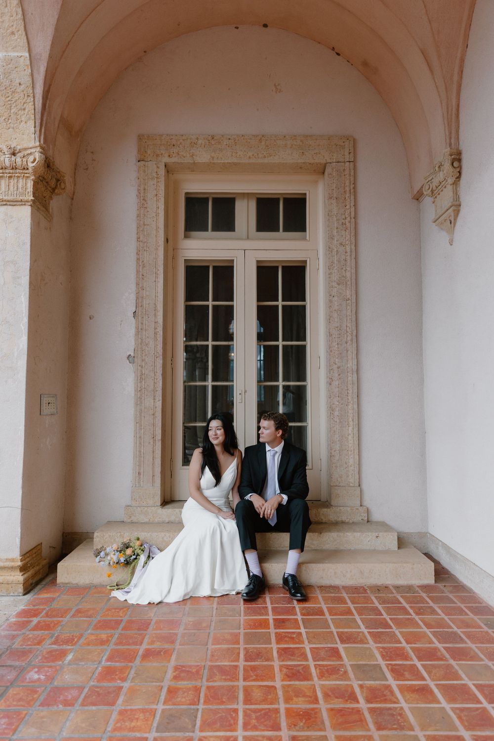 Two people in wedding attire seated on steps under an elegant archway with French doors and terra cotta tiles.
