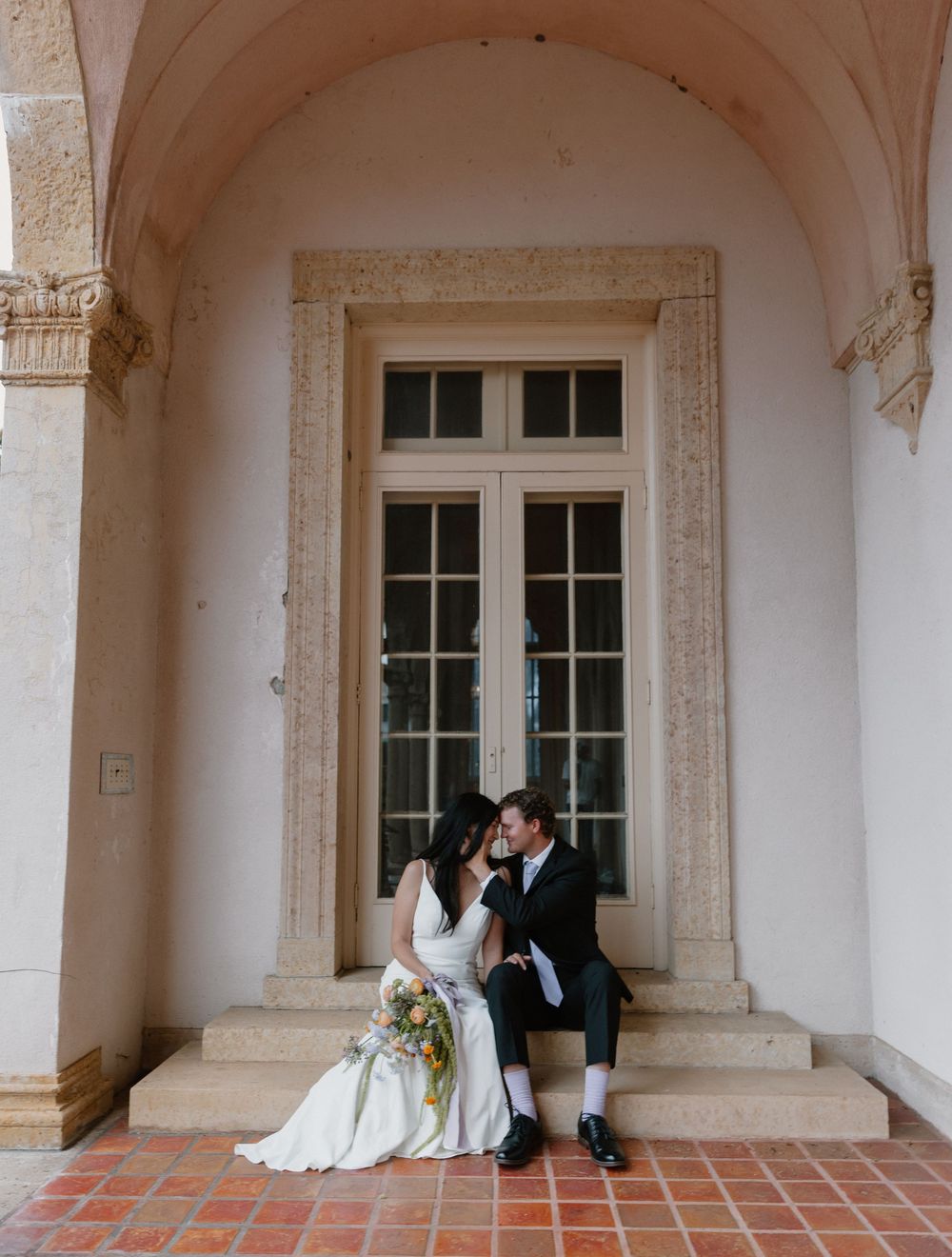 Newlyweds share a tender moment under elegant archways with terracotta tiles and white columns.