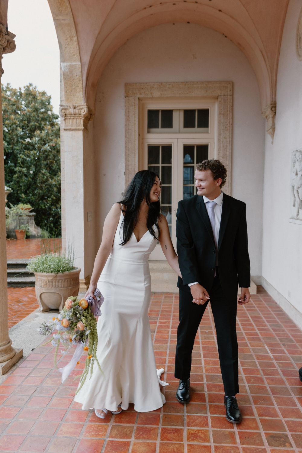 Wedding couple walks playfully through a Mediterranean-style colonnade with brick flooring.