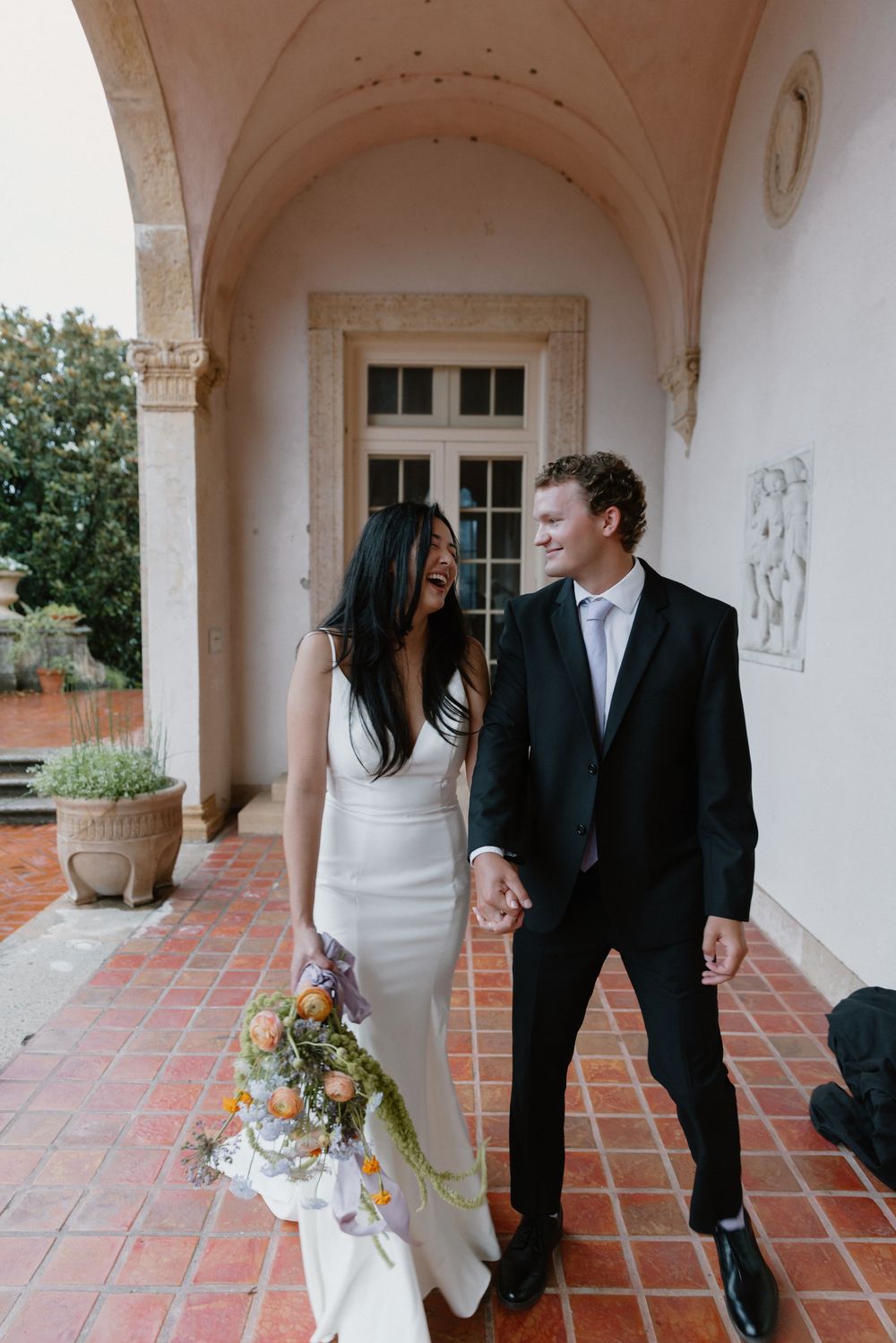 Romantic couple interacts in a light-filled corridor with arched columns and terra cotta floors.