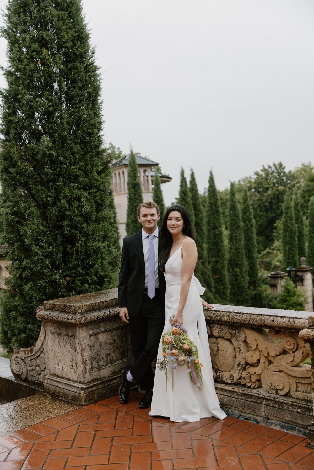 A couple poses on a terrace with stone balustrades and tall cypress trees in an elegant garden setting.