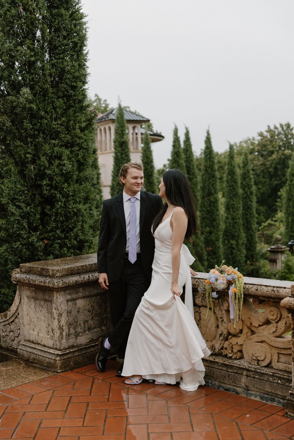 Couple in wedding attire share intimate moments while posing along a rustic brick terrace with cypress trees in the background.