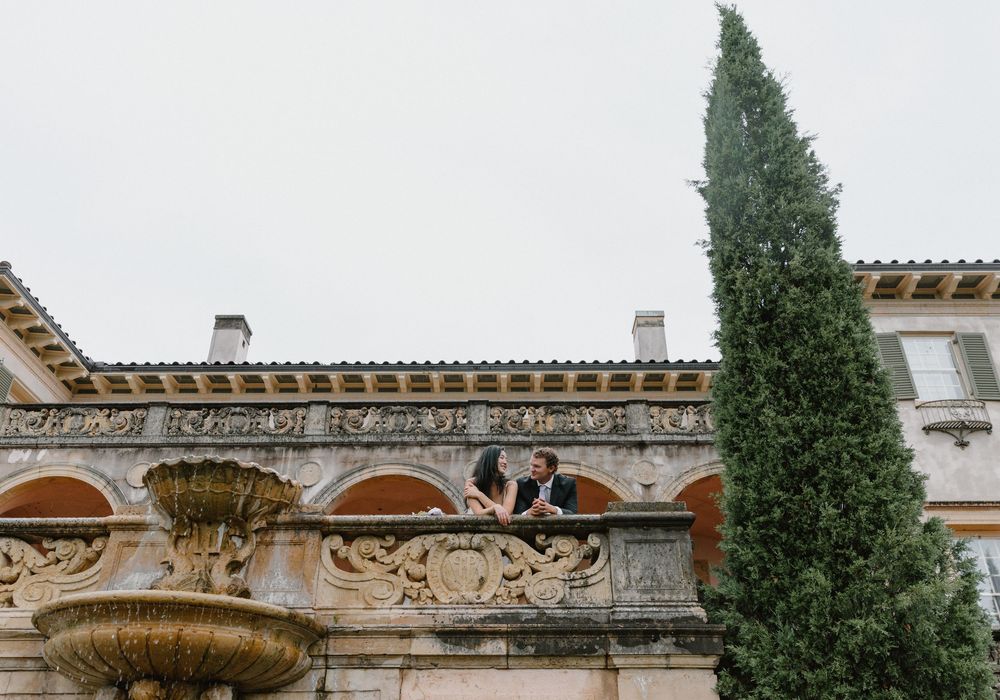 Tall cypress tree stands beside ornate Mediterranean-style villa with decorative stone archways and balustrades.