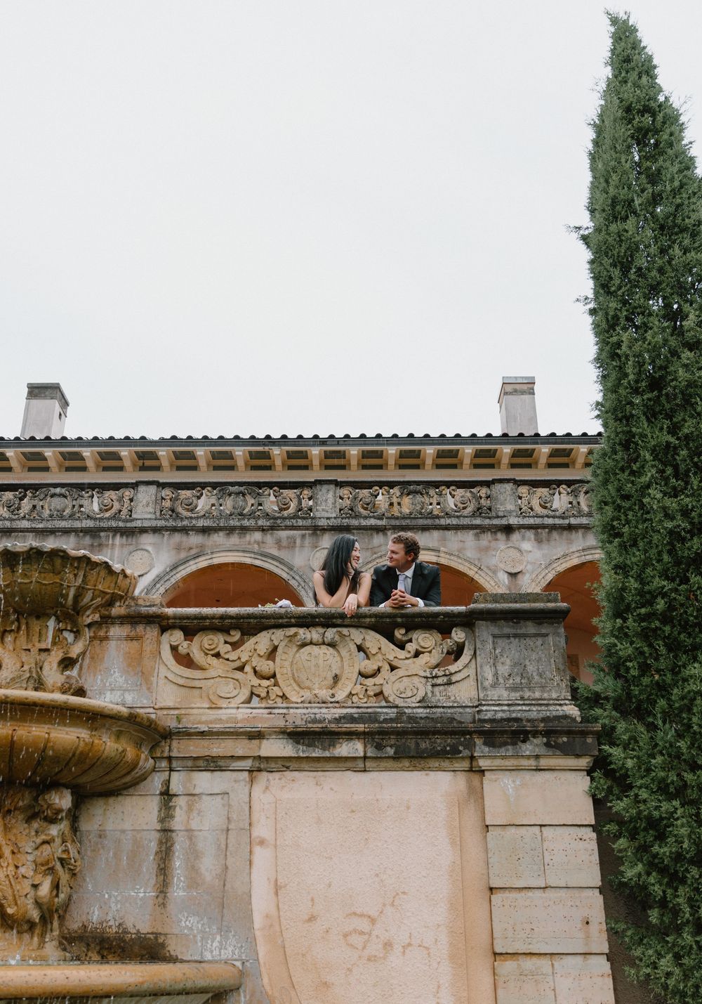 A couple shares an intimate moment atop a Mediterranean stone wall with cypress trees in view.