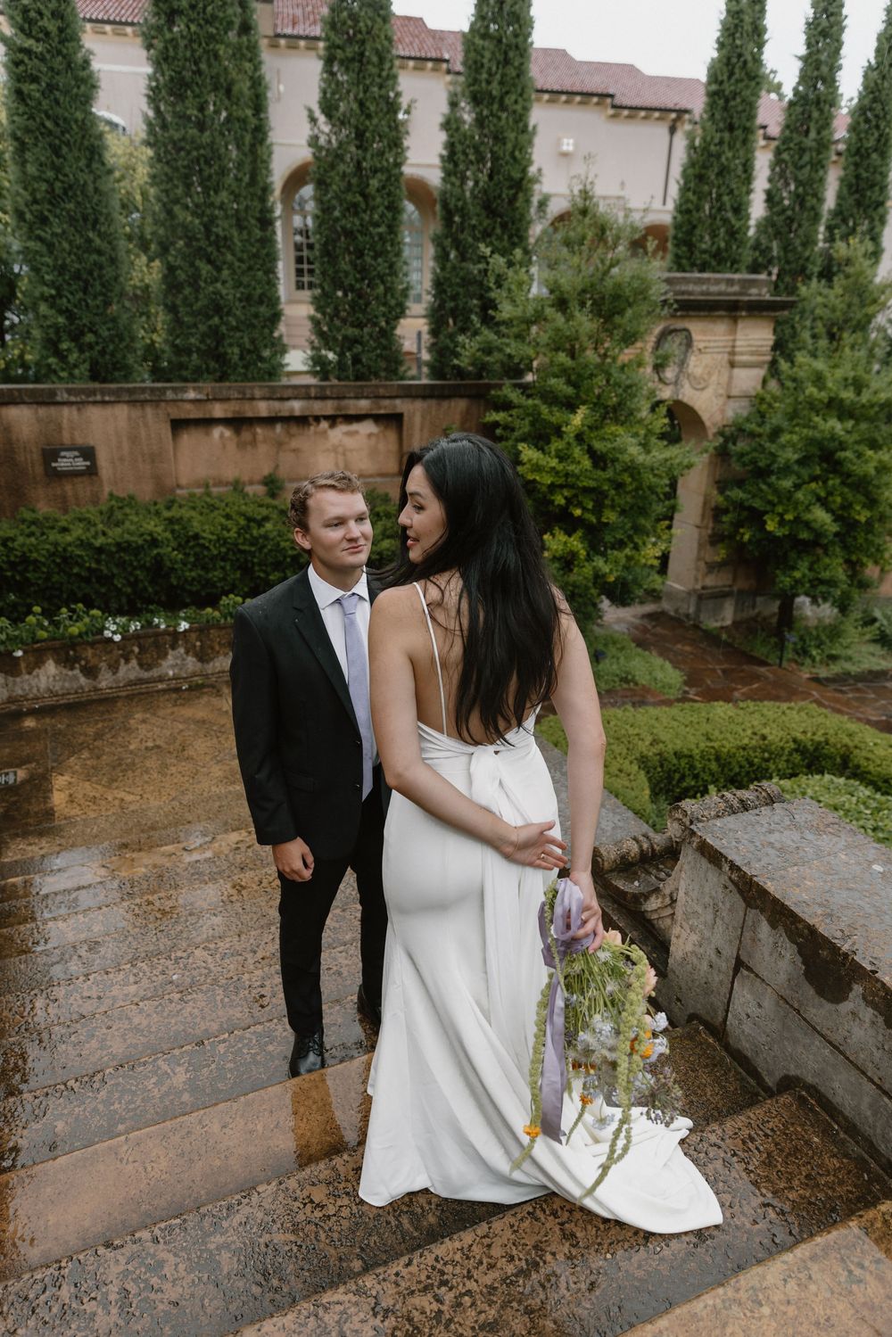 A romantic couple stands together on garden steps surrounded by cypress trees and stone architecture.