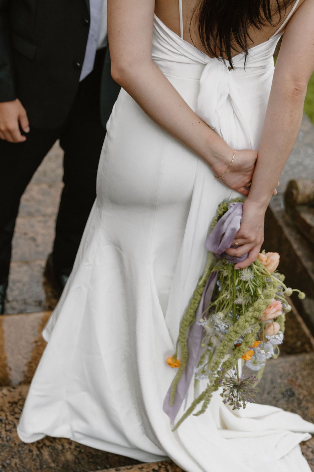 Close-up of a white wedding dress and delicate wildflower bouquet against a stone pathway.