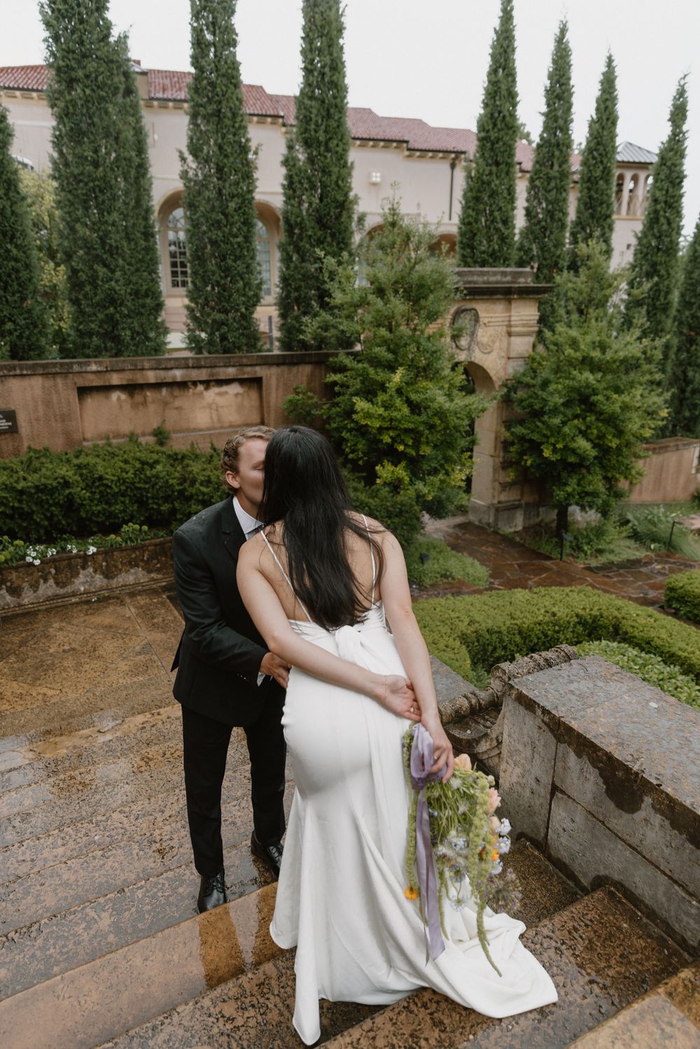 A couple in wedding attire share an intimate moment in a garden courtyard with tall cypress trees and stone walls.